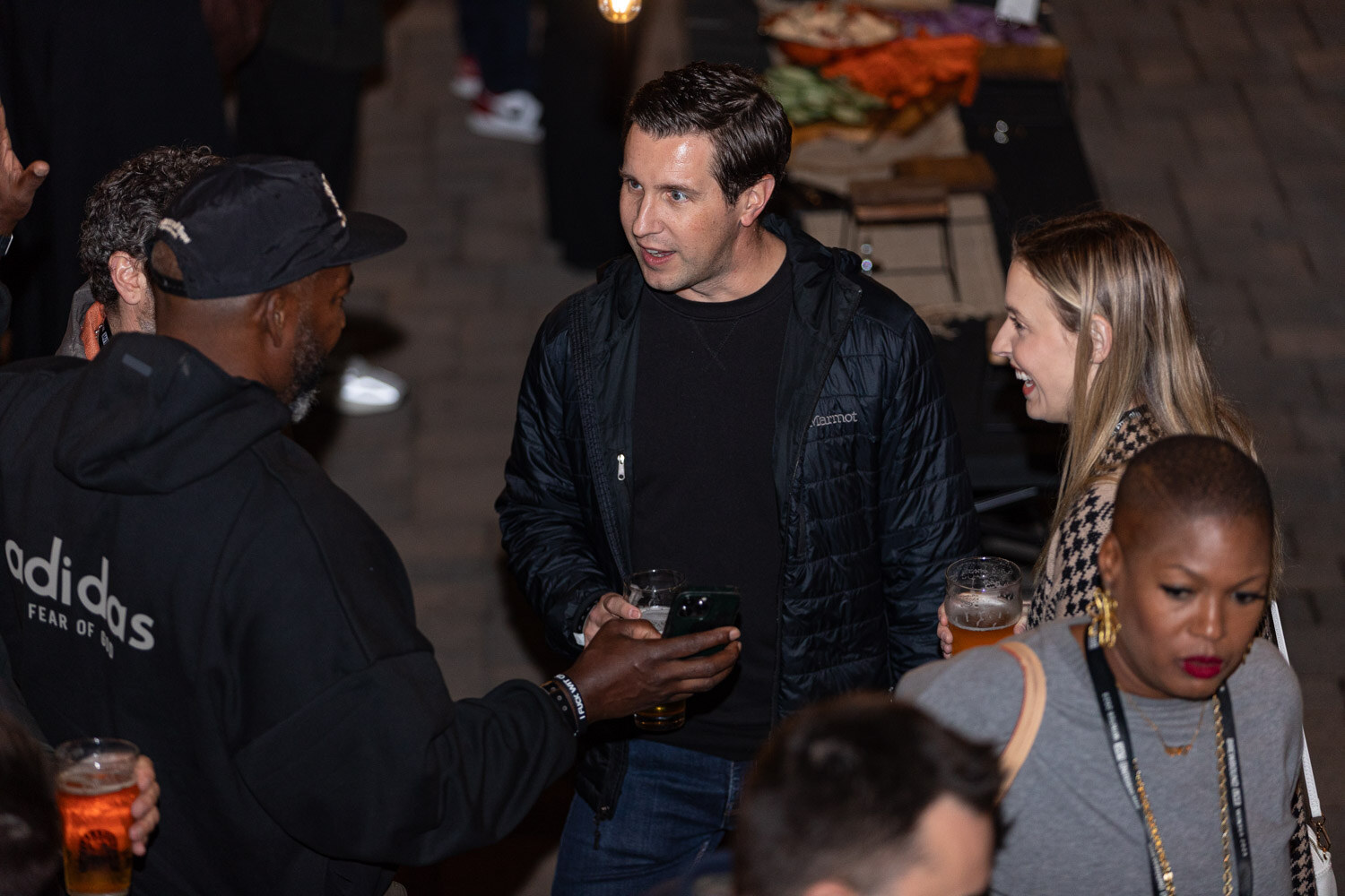 Several guests hold glasses of beer and talk outdoors at night, one in a black adidas hoodie.