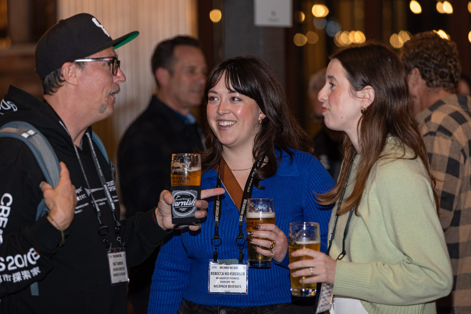 Two women and a man with lanyards smile and hold pints of beer in a crowd.