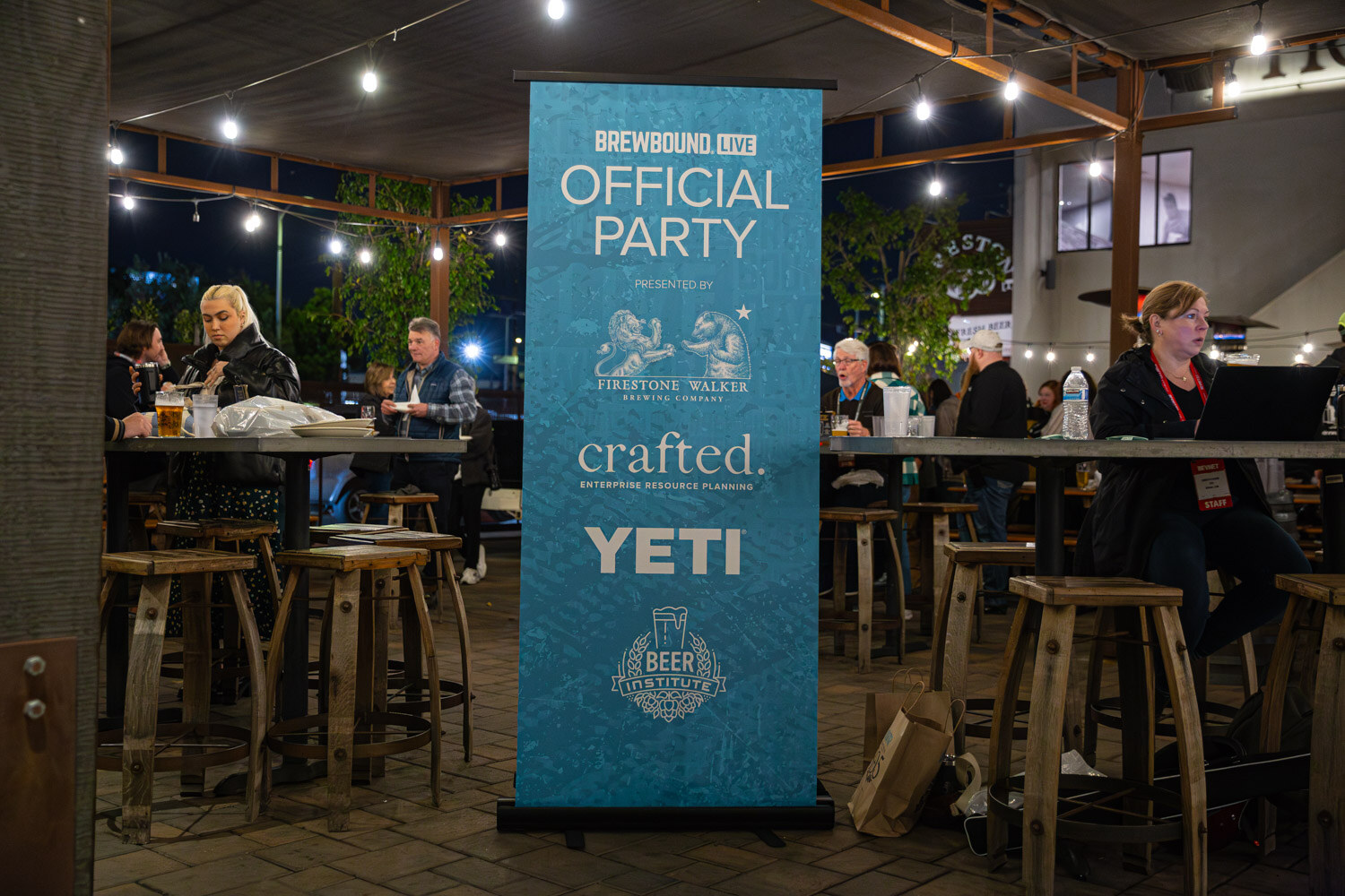 A teal banner reading Brewbound Live Official Party with logos for Firestone Walker, Crafted and YETI stands among bar stools and string lights.