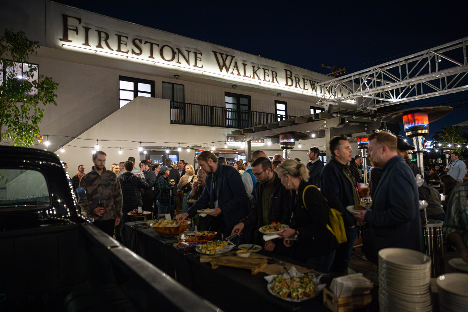 Guests line up at a buffet table next to a vintage truck bed under a Firestone Walker Brewing sign at night.