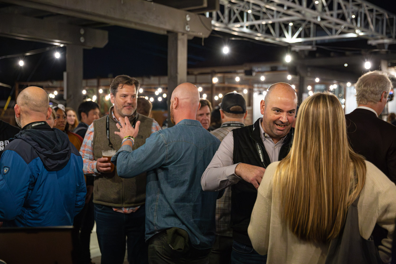 A smiling man in a black vest talks with other guests under string lights on a brewery patio.