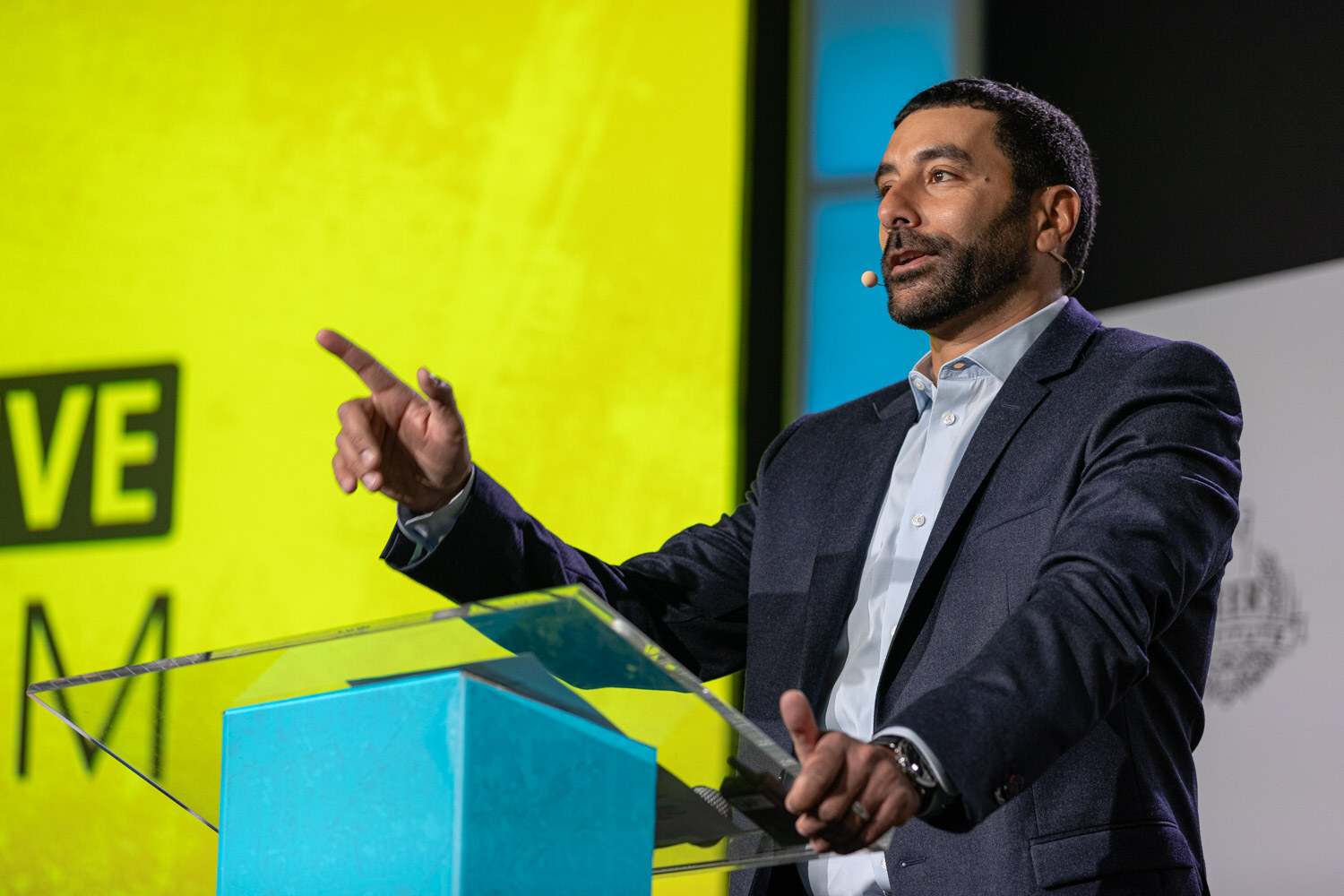 A man in a navy blazer points while speaking at a clear lectern in front of a bright yellow slide.