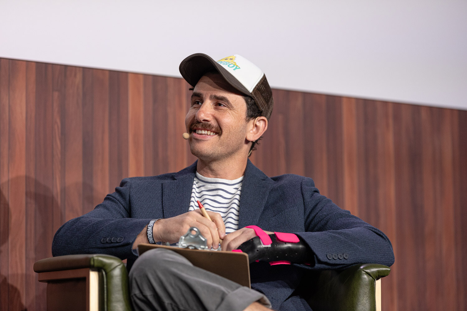A smiling man with a mustache in a trucker cap, striped shirt and navy blazer sits in an armchair holding a clipboard.