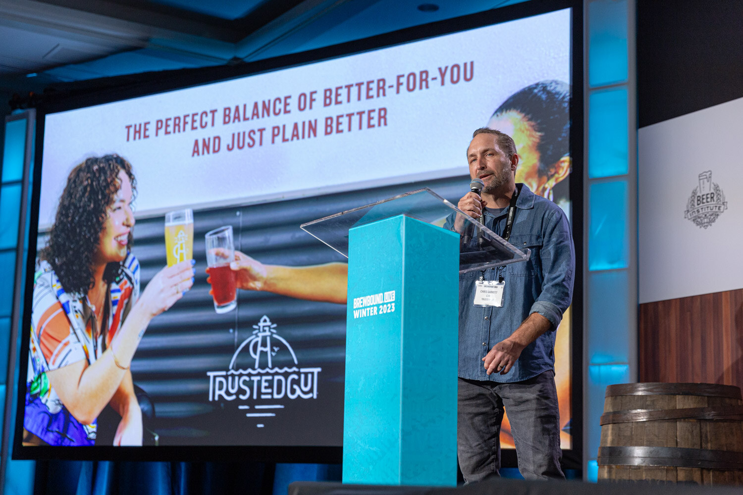 A man in a denim shirt speaks at a lectern beside a large screen showing two people holding drinks and the Trusted Gut logo.