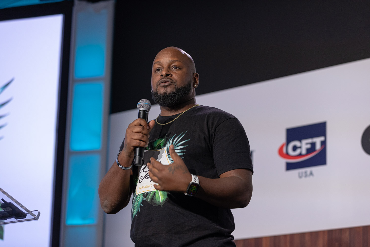 A man in a black graphic t-shirt holds a microphone on stage with a CFT USA logo on the wall behind him.
