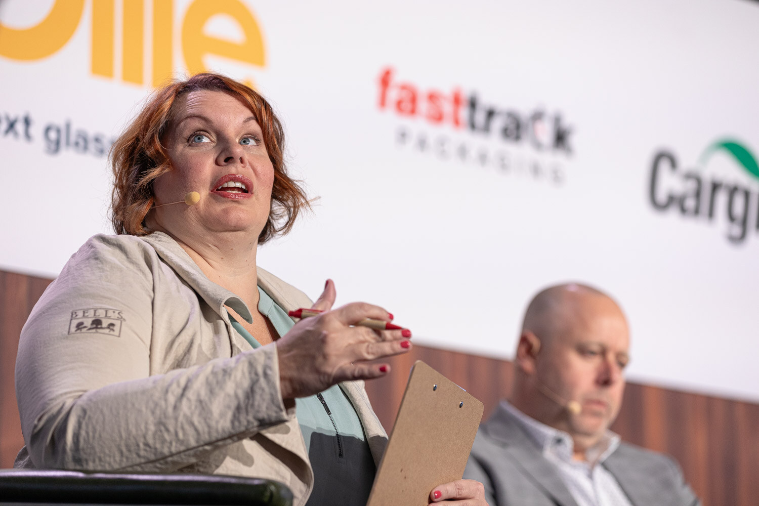 A woman with auburn hair gestures and looks upward while a bald man sits beside her in front of a sponsor banner.