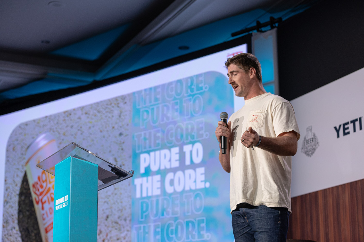 A man in a cream t-shirt and jeans holds a microphone next to a lectern and a screen reading The Core, Pure to the Core.