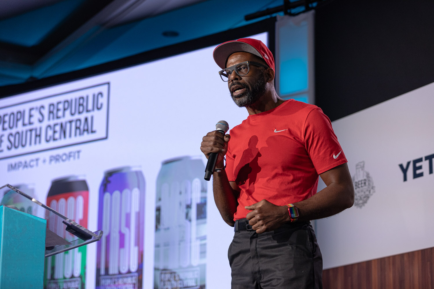 A man in a red t-shirt and red cap holds a microphone in front of a slide reading People's Republic of South Central with three cans.