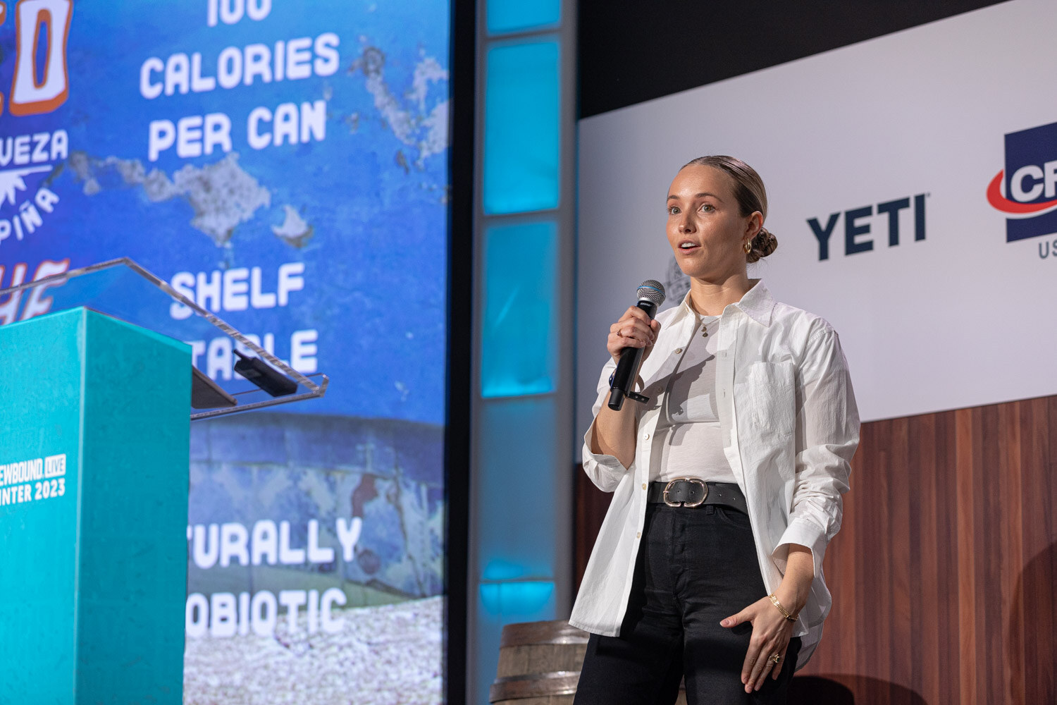 A woman in a white button-up shirt holds a microphone beside a lectern and a blue slide reading 100 Calories Per Can.