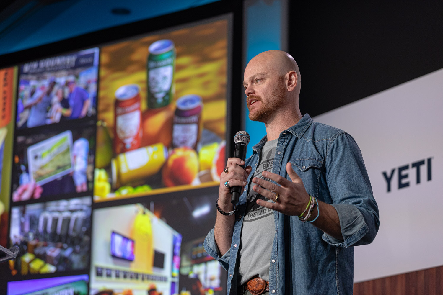 A bald bearded man in a denim shirt holds a microphone in front of a collage slide of cans and product photos.