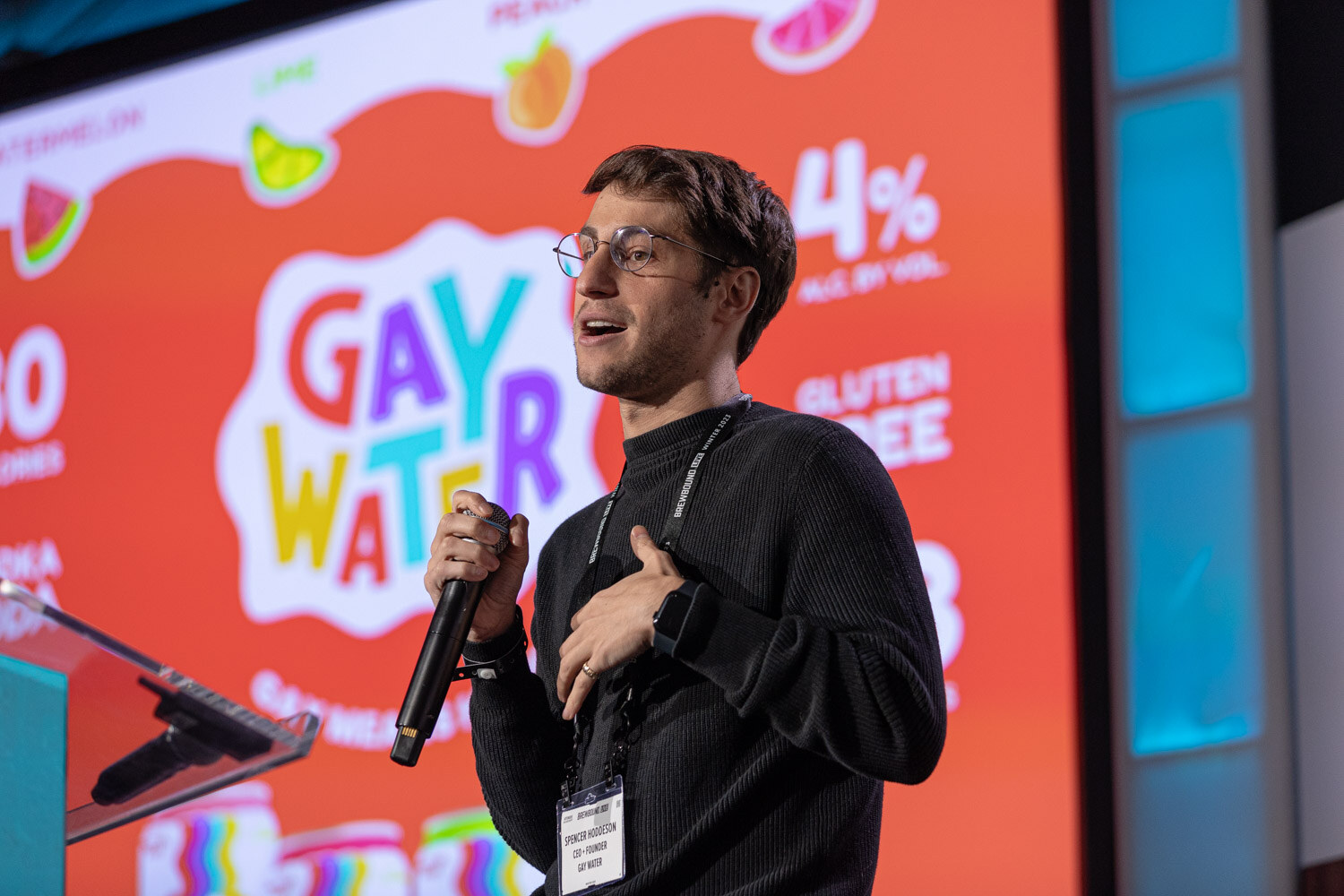 A man in glasses and a black sweater holds a microphone in front of an orange slide with Gay Water lettering.
