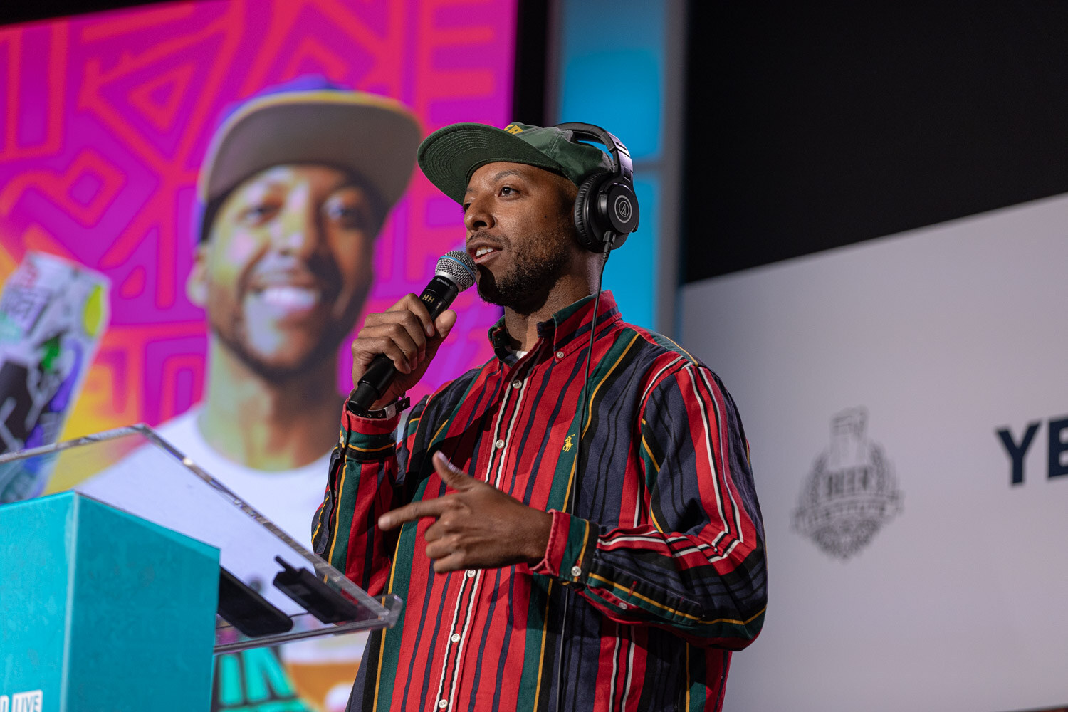 A man in a green cap and headphones around his neck holds a microphone in front of a magenta slide with a smiling face.