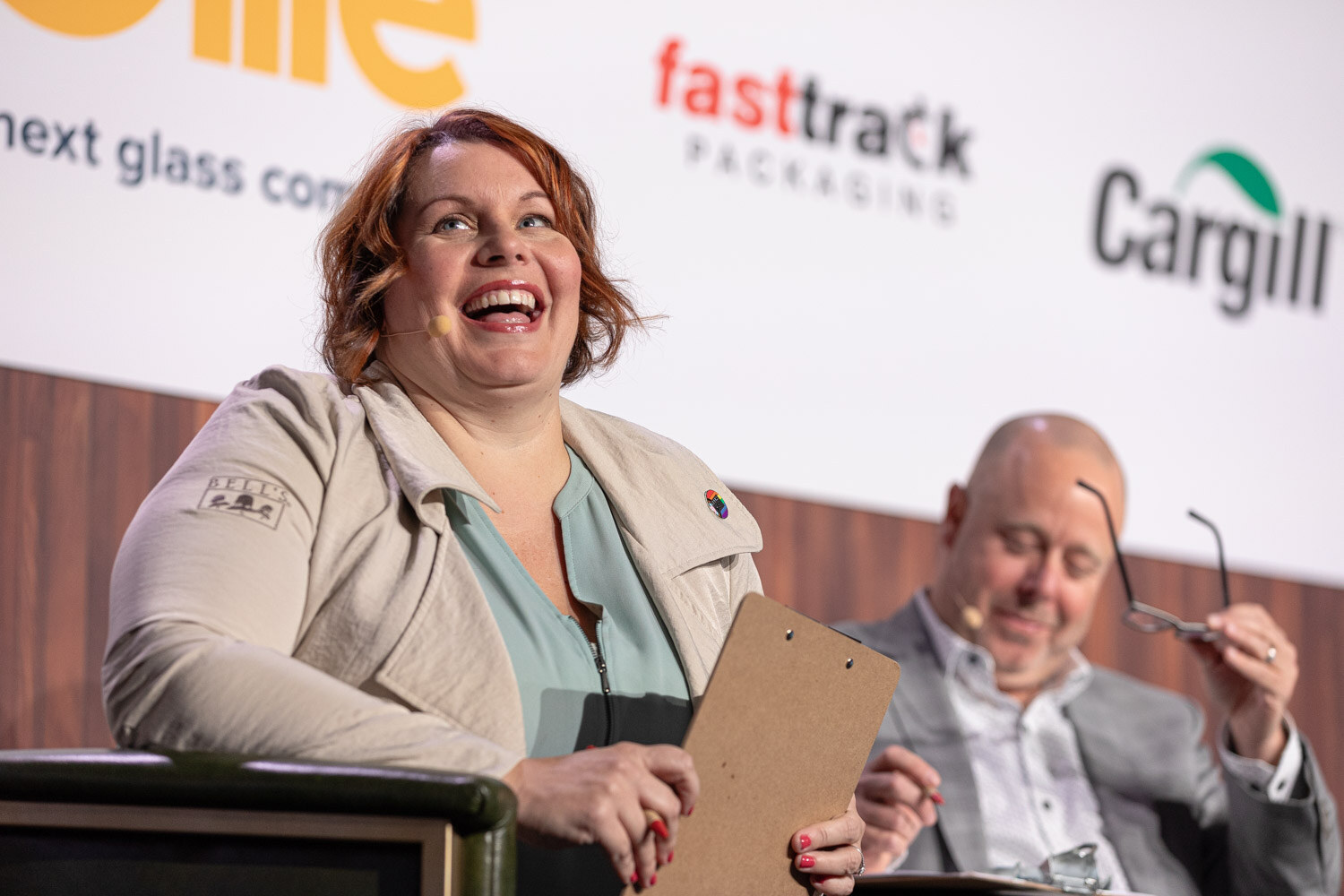 A woman with auburn hair laughs while holding a clipboard as a bald man beside her looks down holding his glasses.
