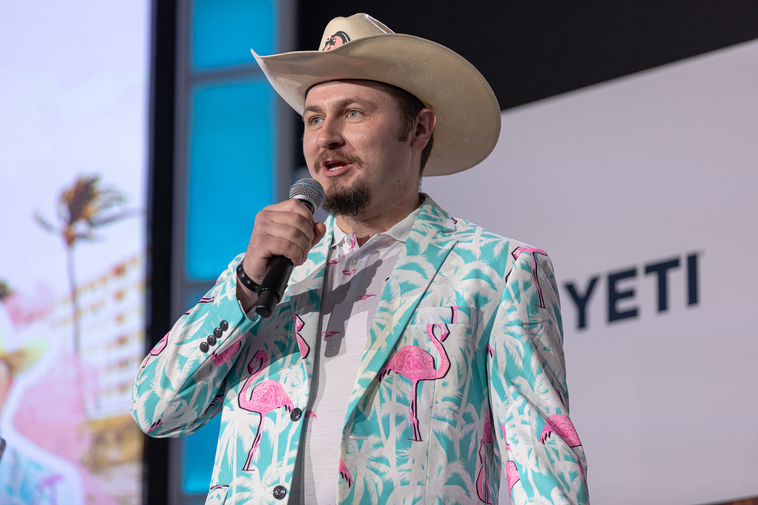 A man in a cowboy hat and a teal blazer printed with pink flamingos holds a microphone.