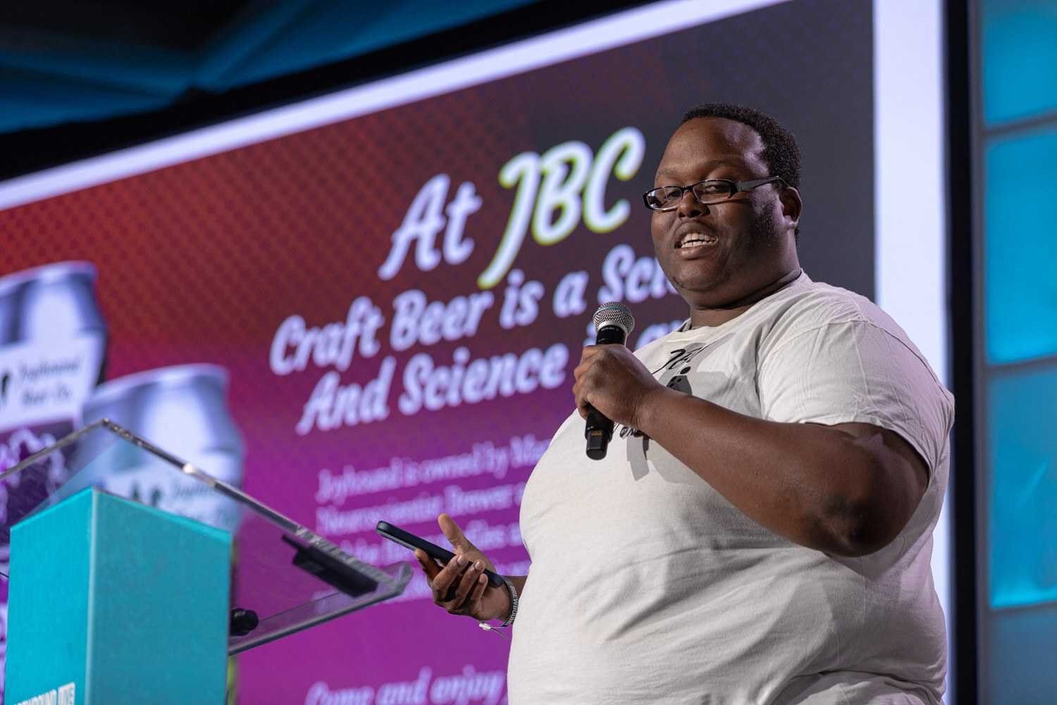 A man in glasses and a white t-shirt holds a microphone in front of a magenta slide reading At JBC Craft Beer is a Science.