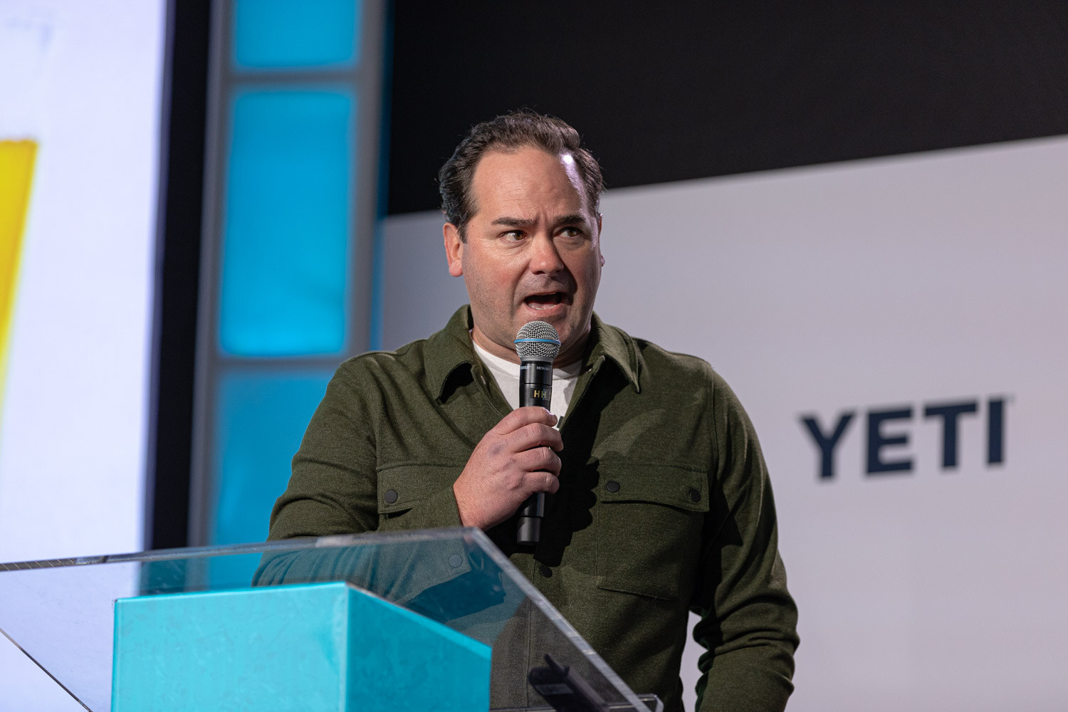 A man in an olive green jacket holds a microphone behind a clear lectern with a YETI banner behind him.