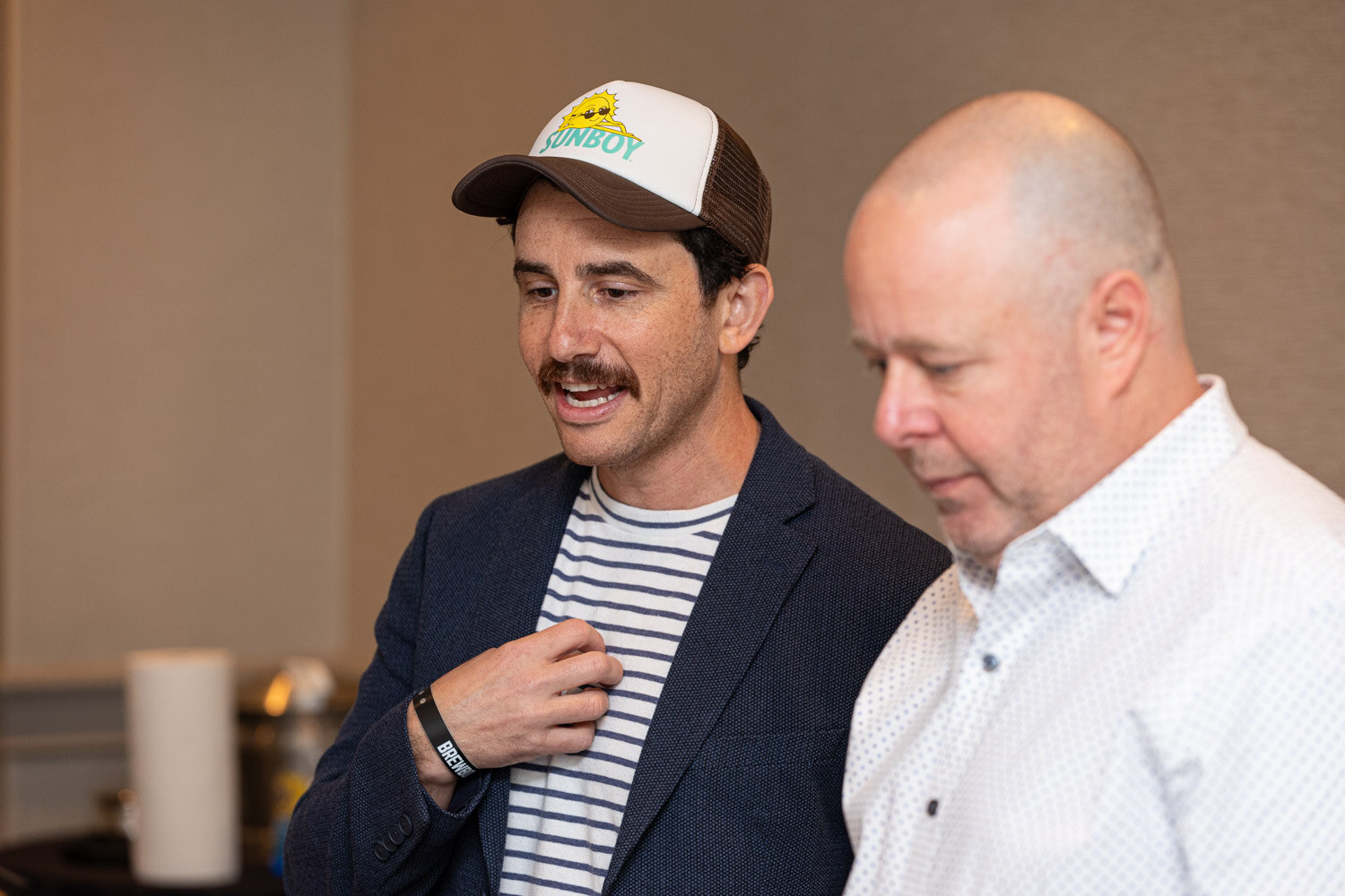 A man in a trucker cap and navy blazer and a bald man in a white shirt look down together in a hallway.