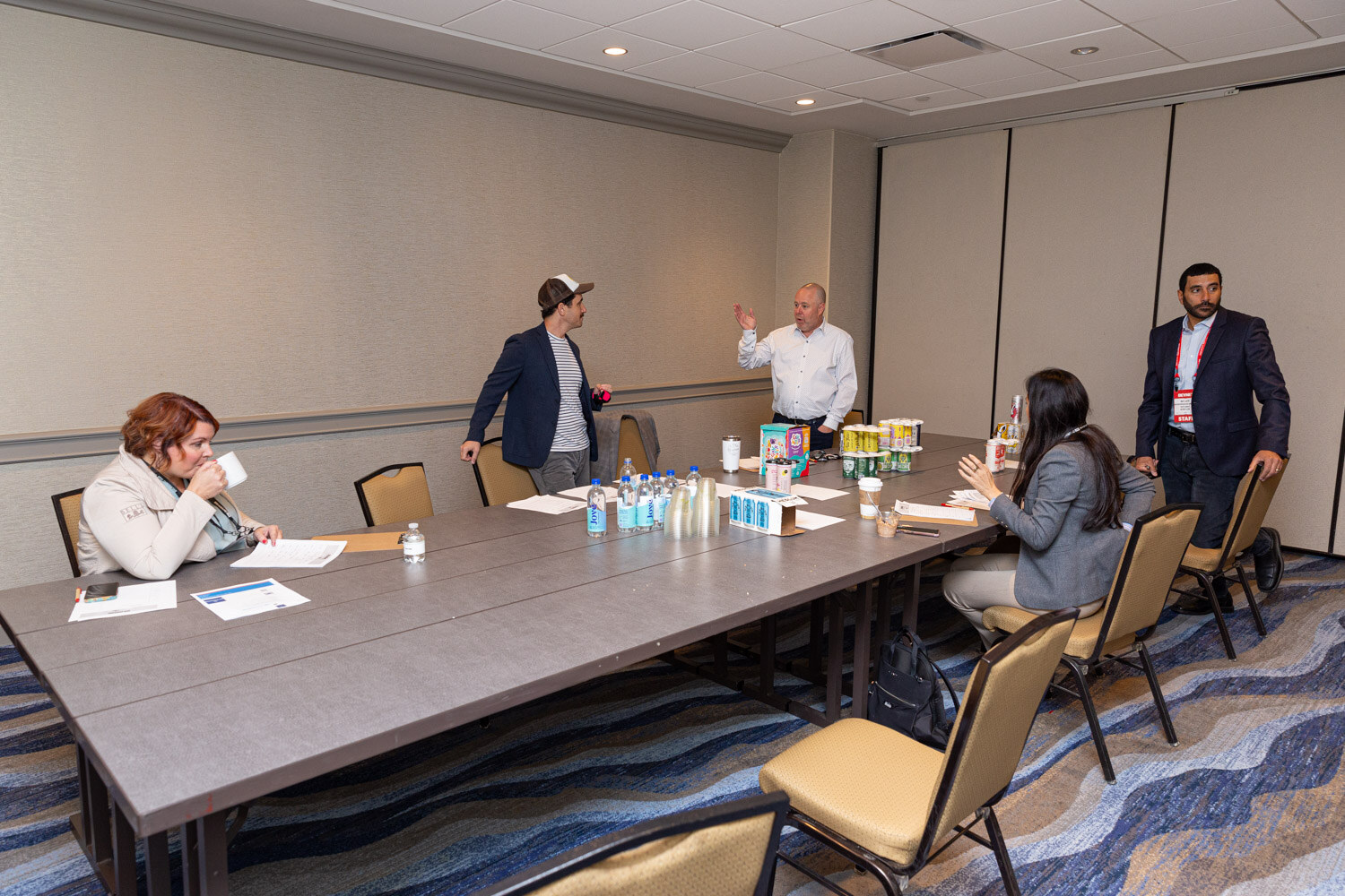 Several people sit and stand around a long conference table with water bottles and beverage cans in a hotel meeting room.