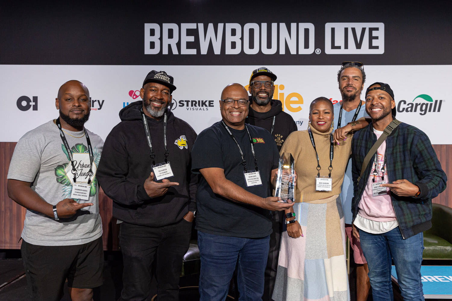 Seven people wearing lanyards pose together on a stage in front of the Brewbound Live backdrop, one holding a glass trophy.