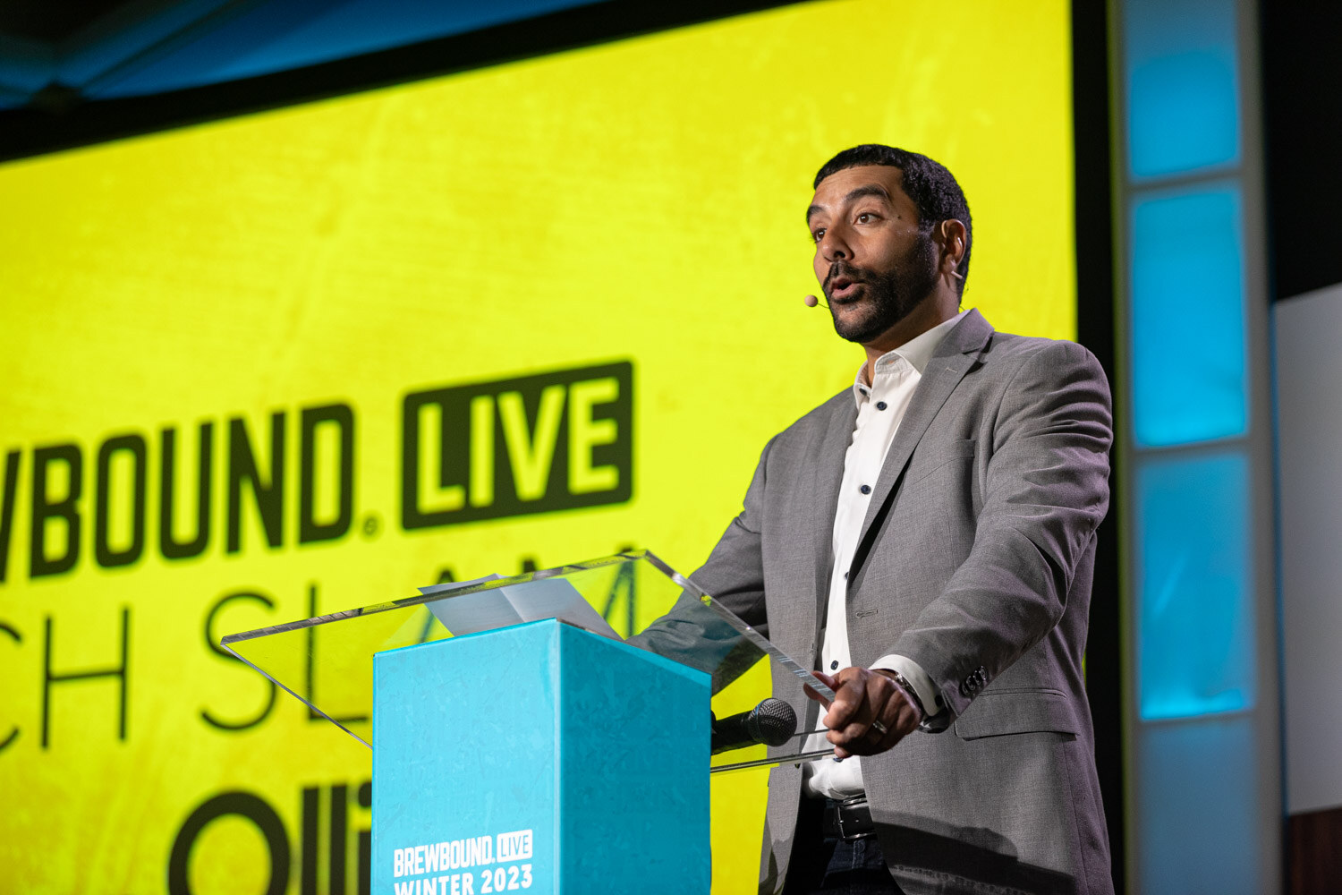 A man in a gray blazer speaks at a clear lectern in front of a yellow screen reading Brewbound Live Pitch Slam.