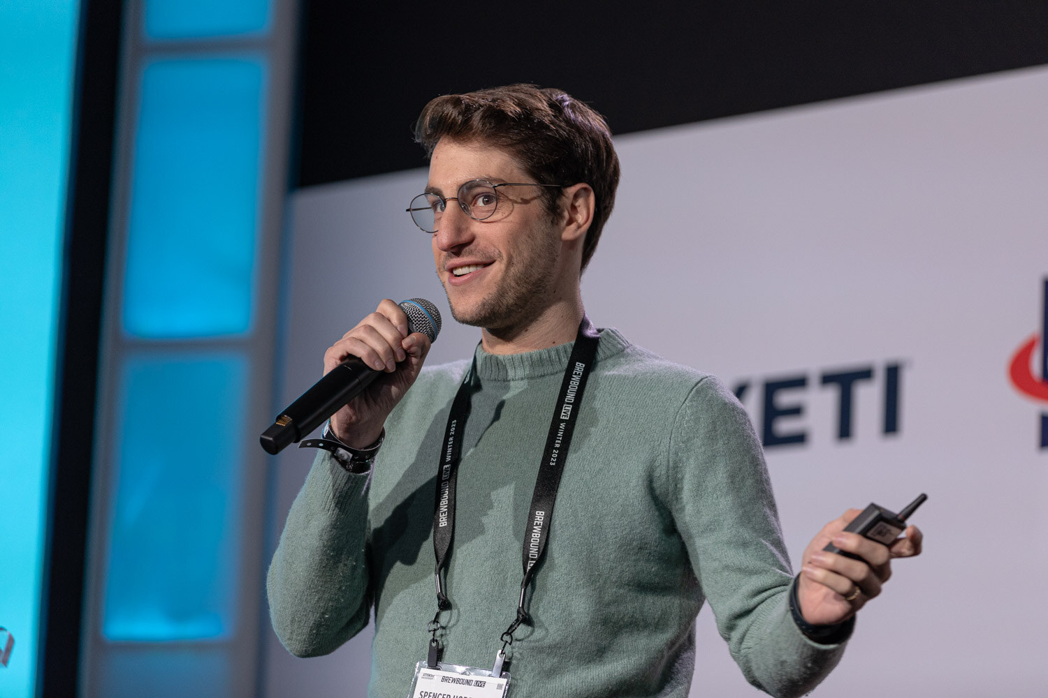 A man in glasses and a green sweater with a lanyard holds a microphone and a clicker in front of a YETI banner.