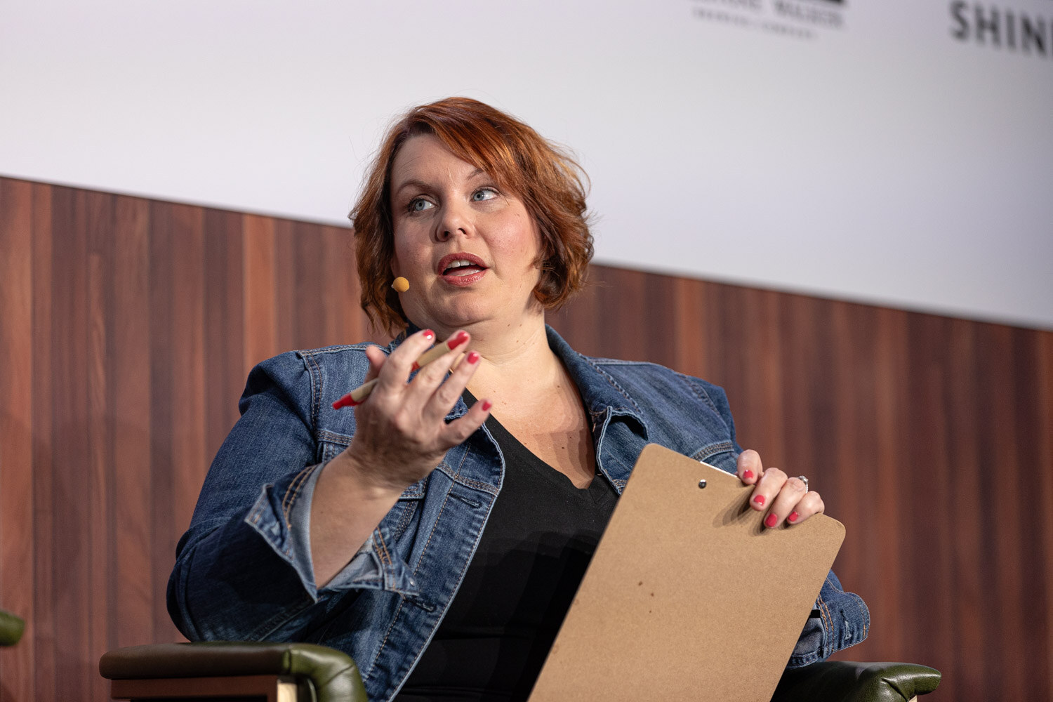 A woman with auburn hair in a denim jacket holds a clipboard and a pen while speaking.