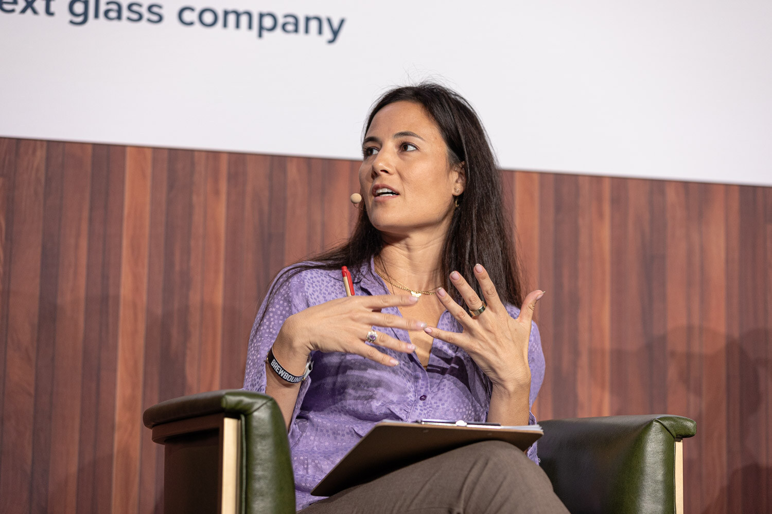 A woman with long dark hair in a lavender blouse sits with a clipboard and gestures with both hands.