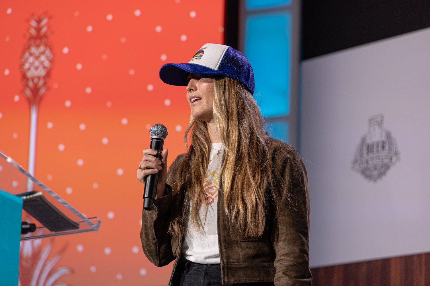 A woman with long hair in a blue trucker cap and brown jacket holds a microphone in front of an orange slide with pineapple art.