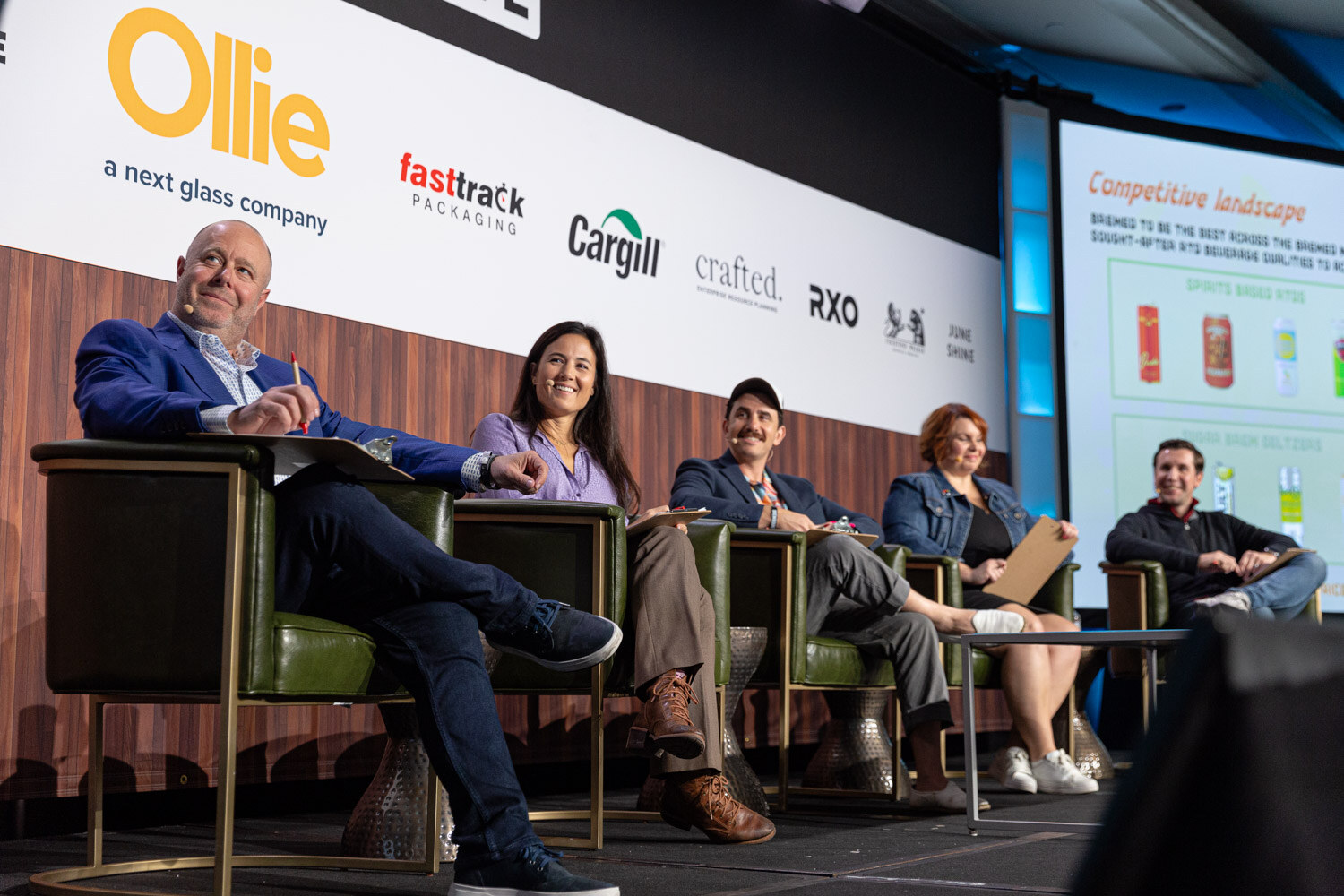 Five people sit in a line of green armchairs on stage next to a screen titled Competitive Landscape.