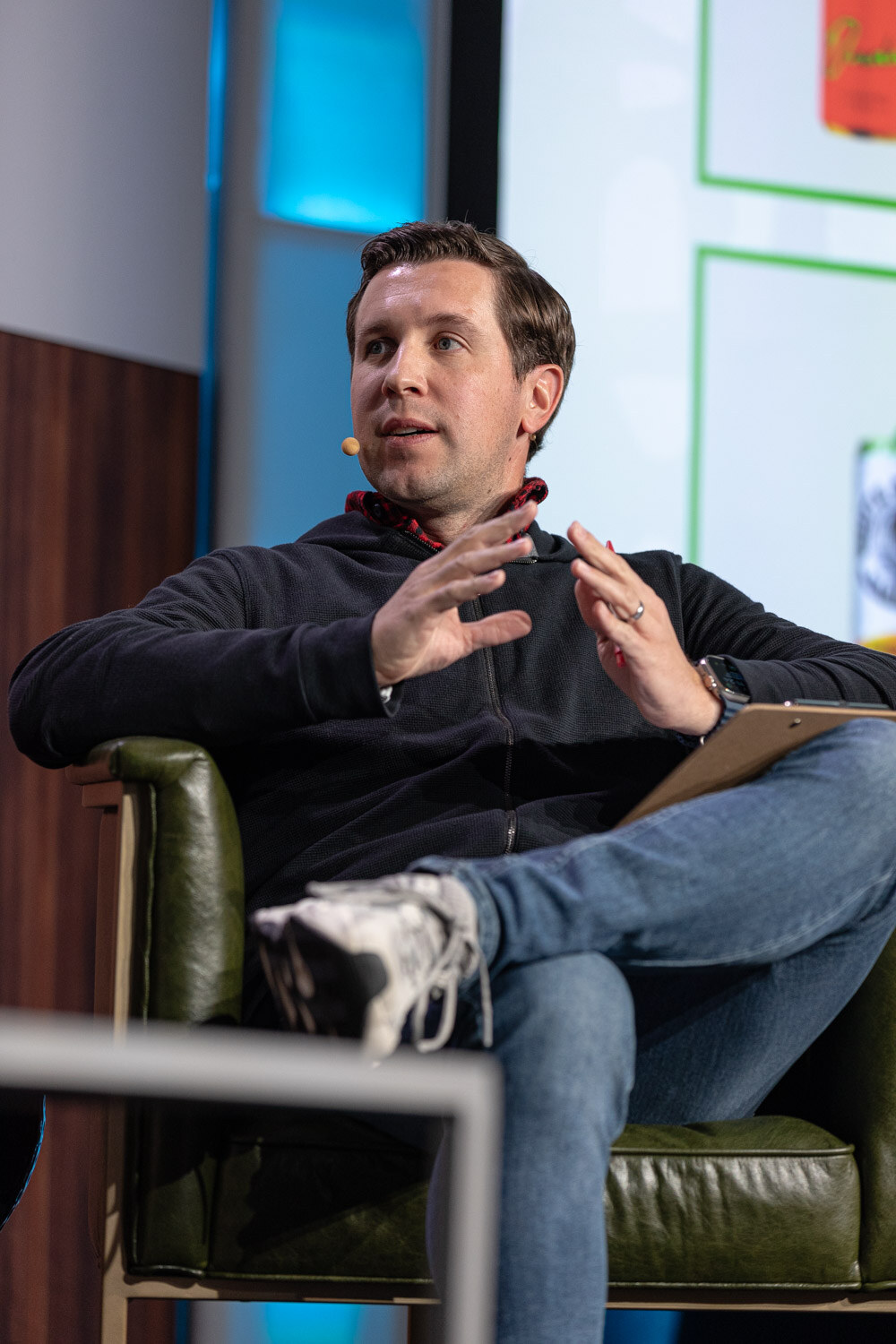 A man in a dark pullover and jeans sits in an armchair with a clipboard on his knee and gestures with both hands.
