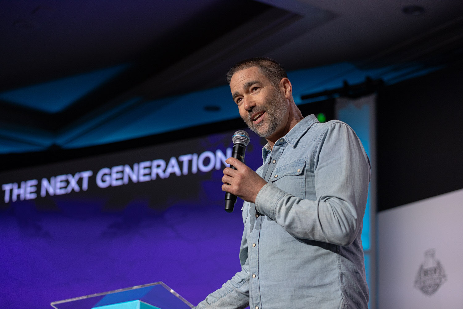 A man in a light denim shirt holds a microphone in front of a purple slide reading The Next Generation.