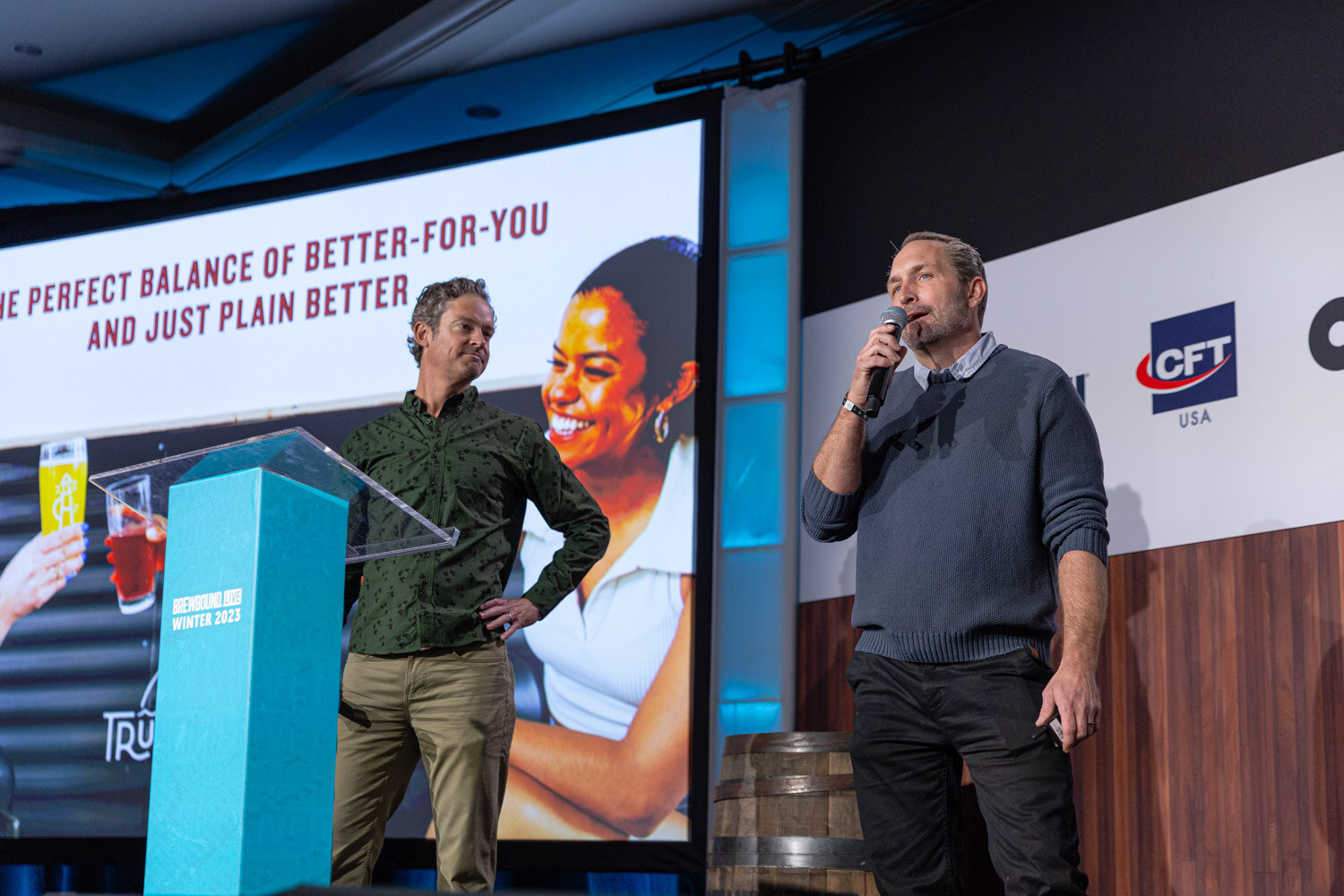 Two men stand on stage on either side of a teal lectern in front of a slide reading The Perfect Balance of Better-for-You.