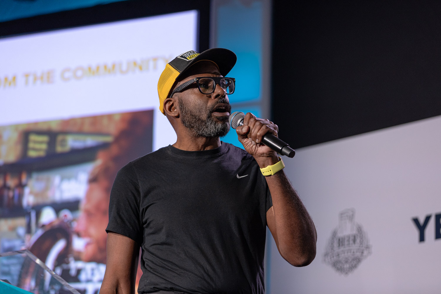 A bearded man in glasses and a yellow cap holds a microphone in front of a slide on stage.