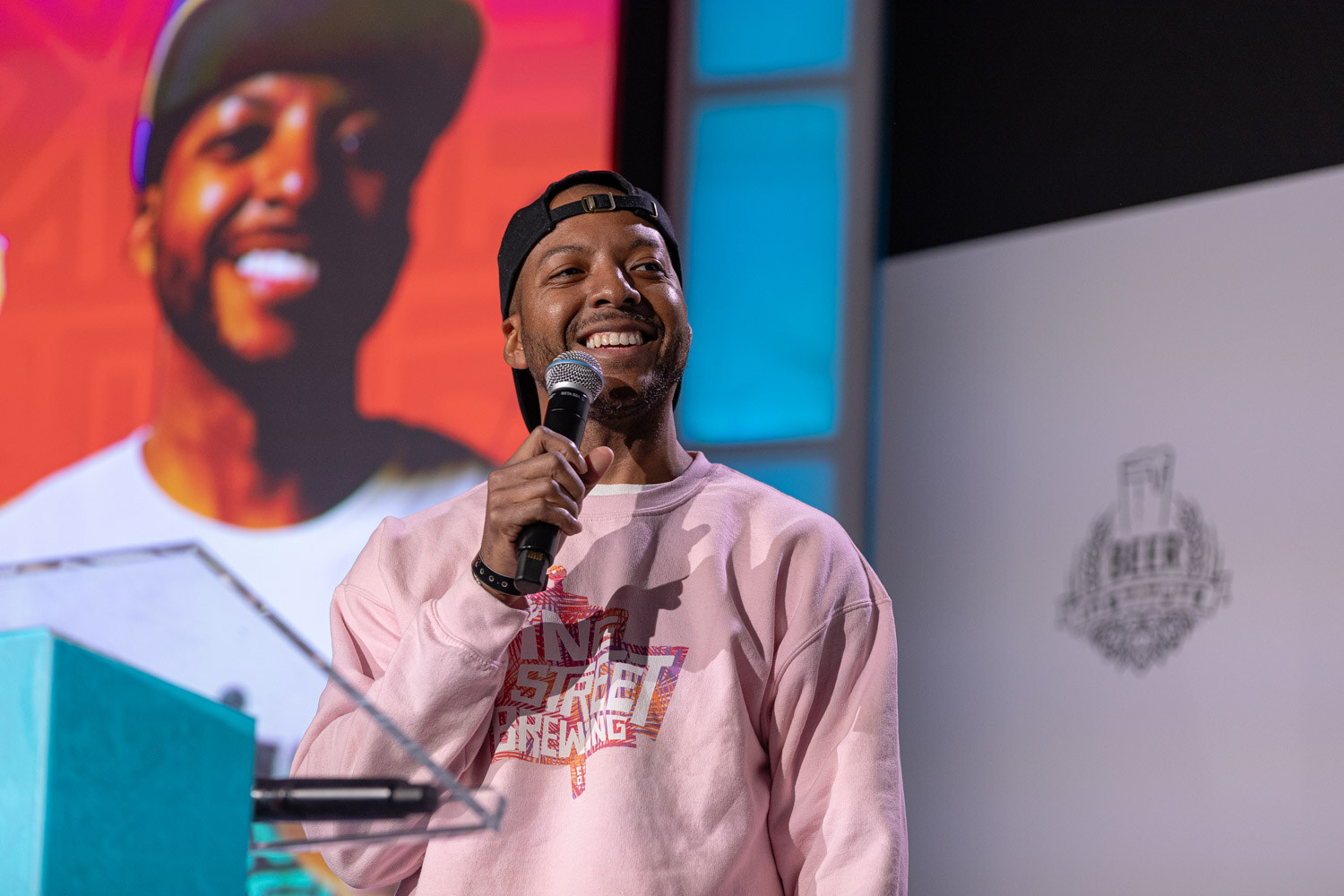 A smiling man in a cap and pink sweatshirt holds a microphone in front of a slide with a large smiling face.