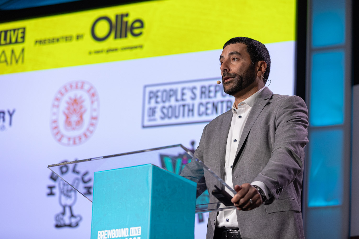 A man in a gray blazer stands at a teal lectern in front of a yellow Pitch Slam screen.