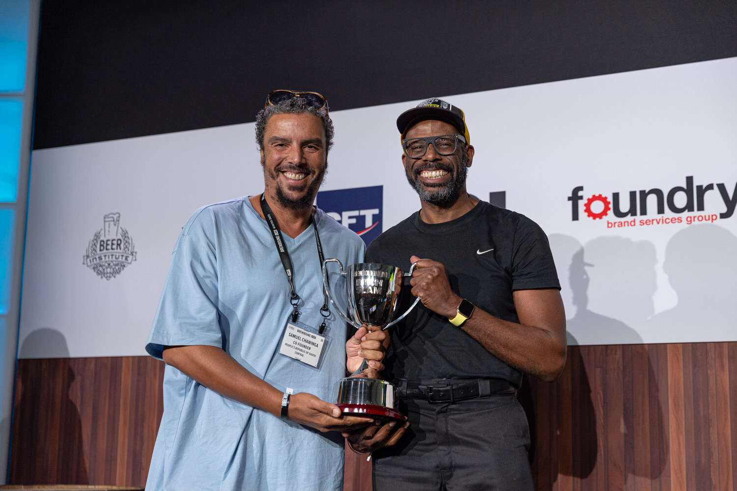 Two smiling men stand together holding a large silver trophy, one in a light blue shirt and one in a black shirt and cap.