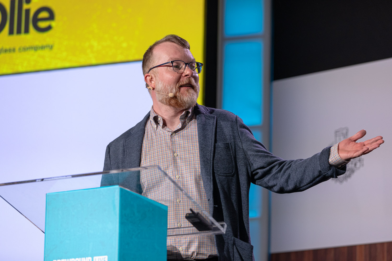 A bearded man in glasses and a gray jacket gestures with one arm outstretched at a clear lectern.
