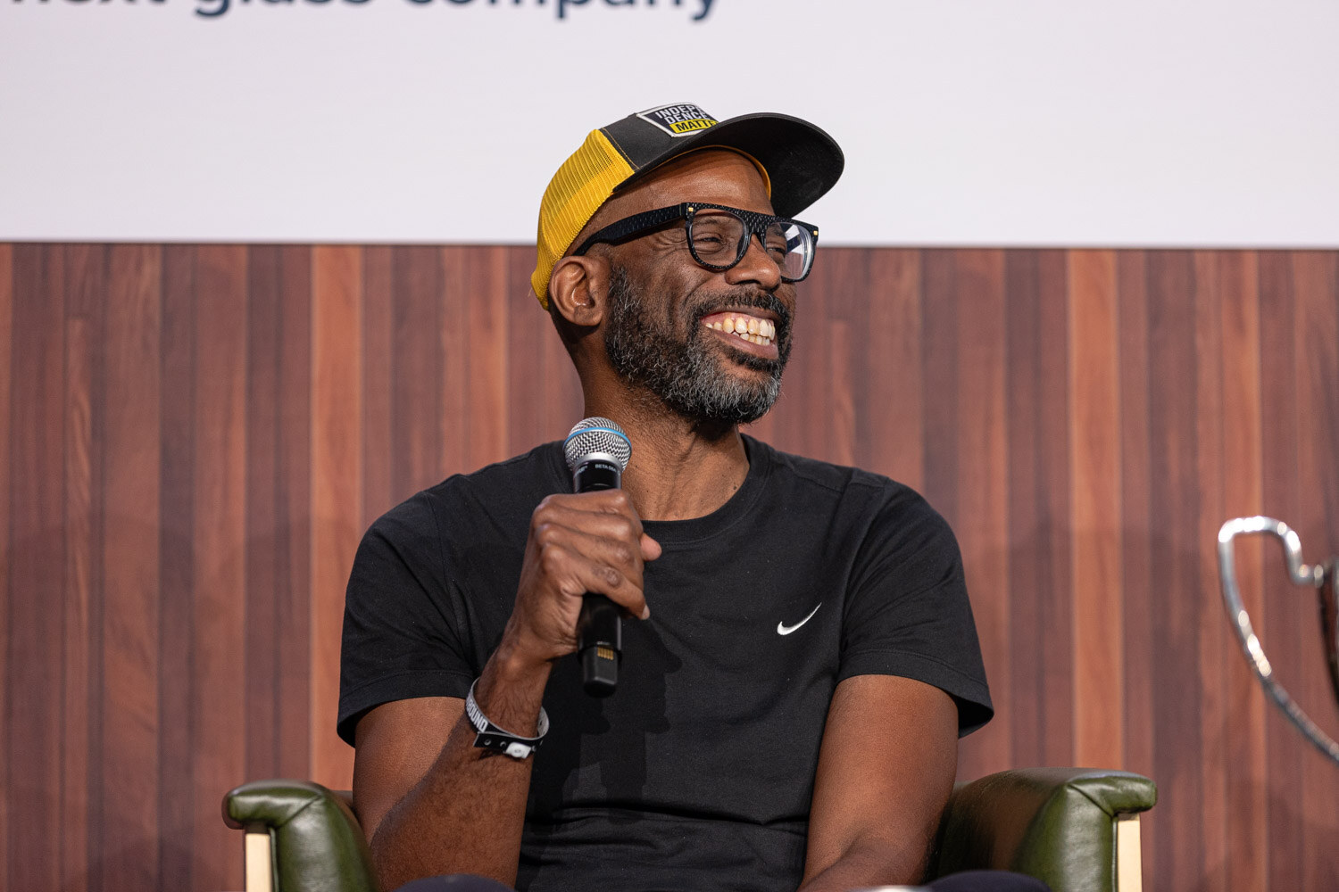 A smiling bearded man in glasses and a yellow cap holds a microphone and laughs.