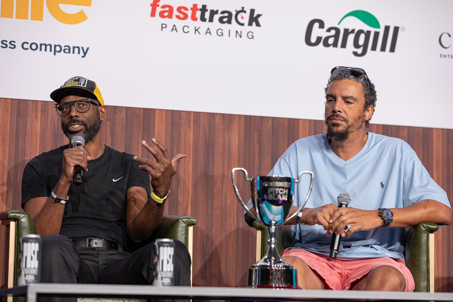 A man in a black t-shirt gestures while speaking, seated beside a man in a light blue shirt with a silver Pitch Slam trophy on the table.
