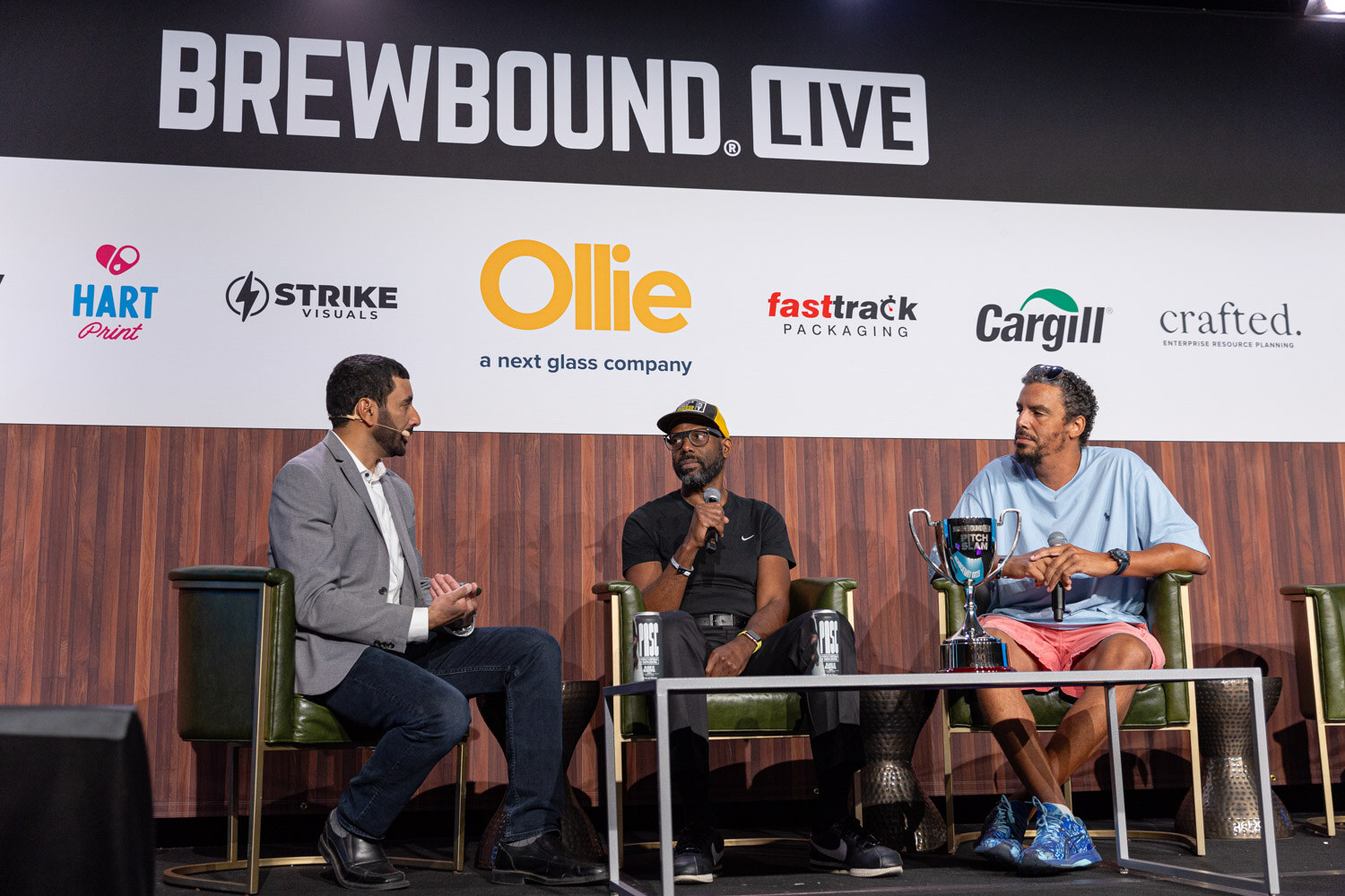 Three men sit on stage in armchairs around a table, one holding a trophy, with the Brewbound Live backdrop behind.