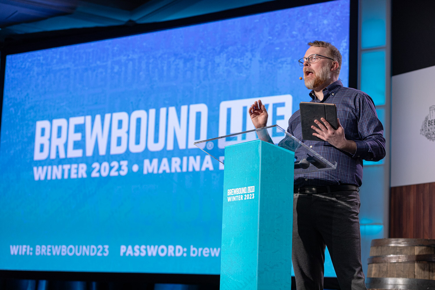 A man in glasses speaks at a teal lectern holding a tablet, with a large blue Brewbound Live Winter 2023 Marina del Rey screen behind him.