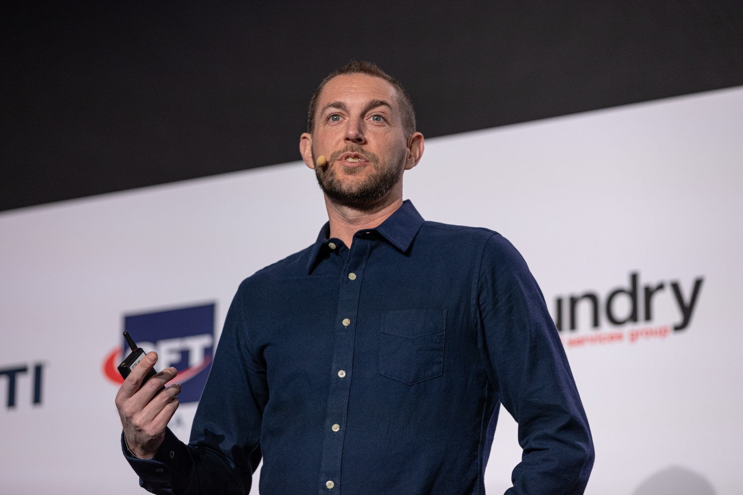 A bearded man in a navy button-up shirt speaks to the room, holding a presentation clicker, in front of a sponsor banner.