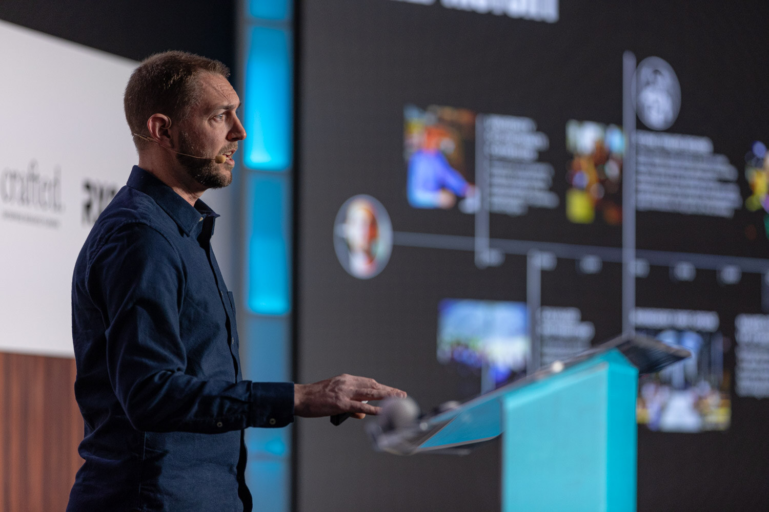 A man in a dark shirt with a headset microphone stands at a lectern beside a large screen showing a timeline slide.