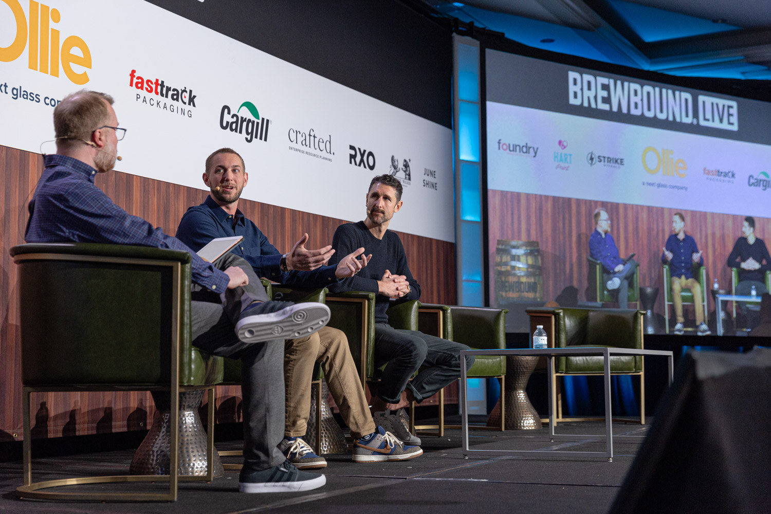 Three men sit in green armchairs on a stage in front of a Brewbound Live backdrop and a screen showing the same stage.