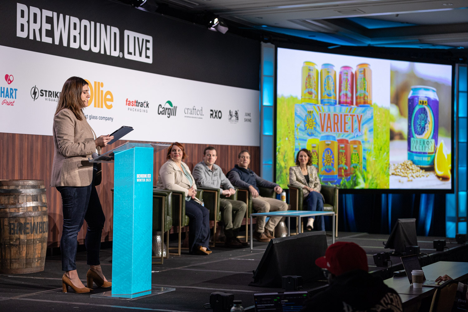 A woman stands at a teal lectern next to four seated panelists, with a screen showing a variety pack of beer cans.