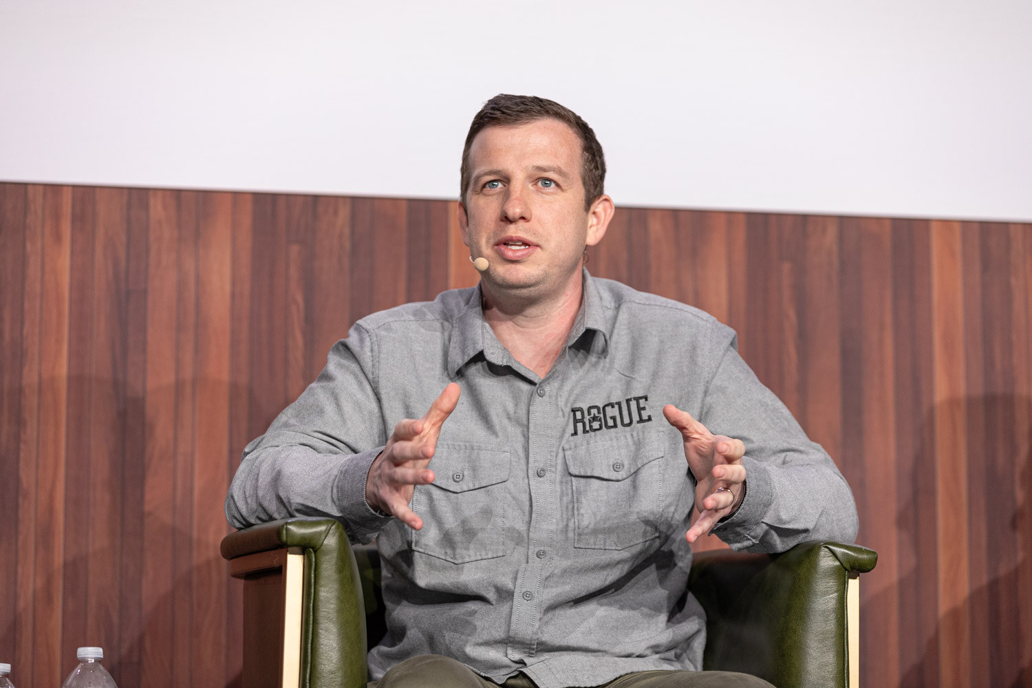 A man in a gray shirt with a Rogue logo sits in an armchair and gestures with both hands while speaking.
