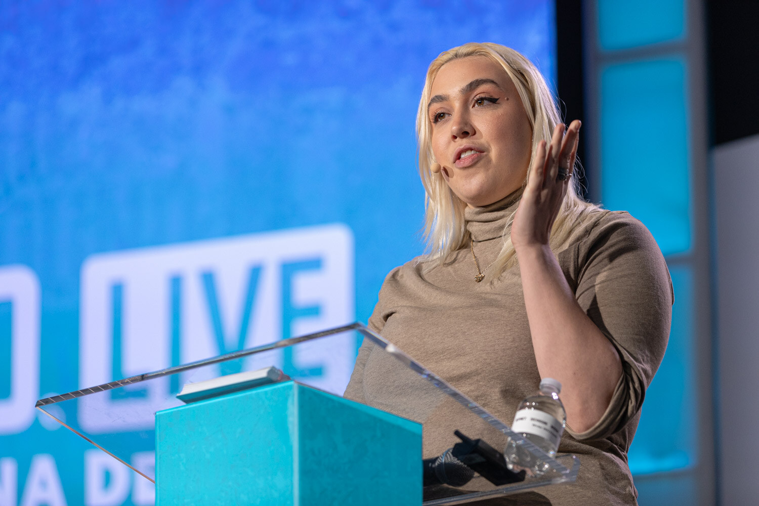 A woman with blonde hair gestures as she speaks at a clear acrylic lectern in front of a large blue Brewbound Live screen.