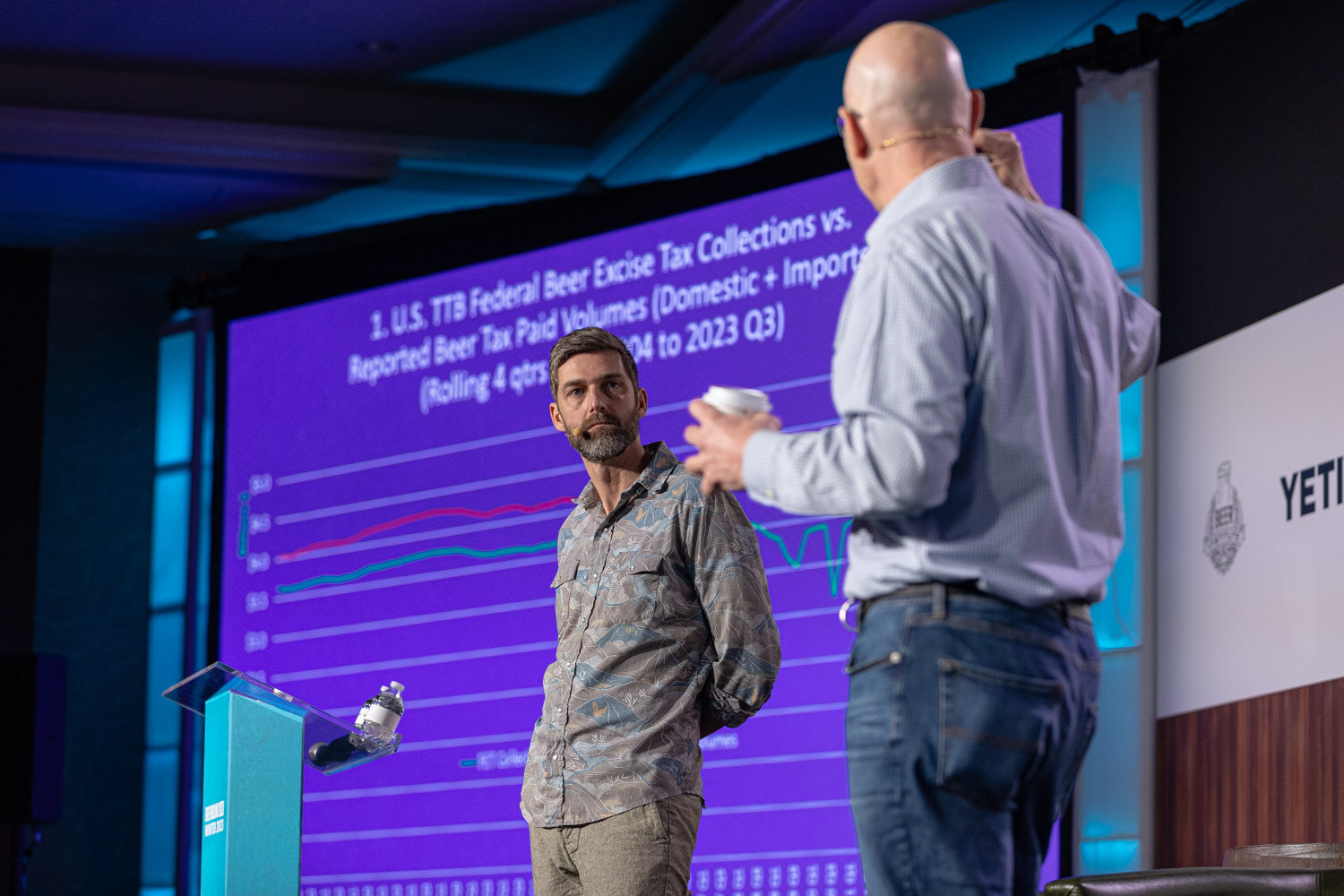 Two men stand on stage in front of a purple slide titled with U.S. TTB Federal Beer Excise Tax Collections and a line chart.