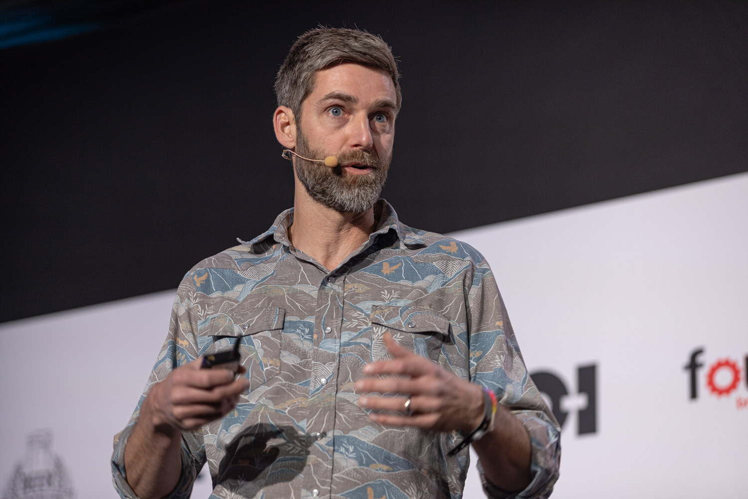 A bearded man in a gray patterned short-sleeve shirt holds a clicker in one hand and gestures with the other in front of a sponsor banner.