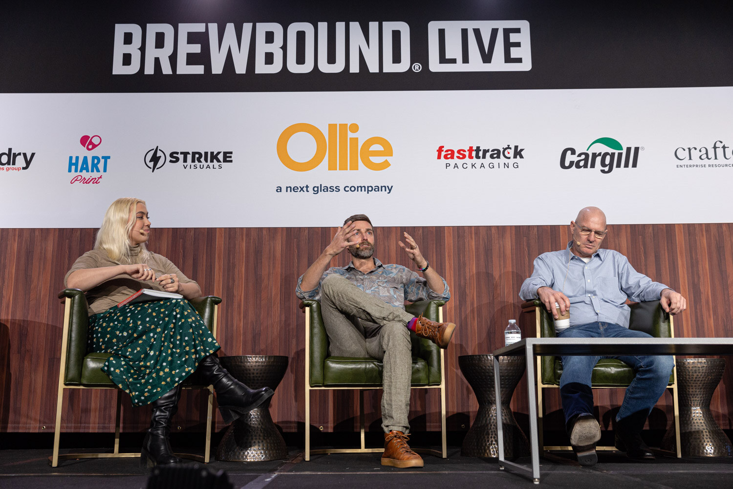 A woman and two men sit in green armchairs on stage in front of a Brewbound Live sponsor banner.